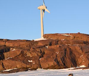 a man sits on the ice holding a camera to his eye. A wind turbine is in the background