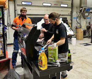 three men stand in front of a bbq in a workshop