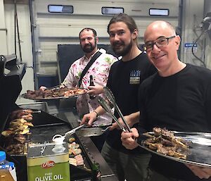 three men stand holding platters in front of a BBQ