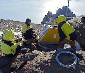 Three men in snow suits discuss how to secure a radio transmitter to the mountain