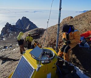 A man in a snow suit is repairing a radio transmitter. Two men stand in the distance with a mountain peak and plateau behind them