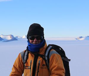 A man in a yellow snow suits stands on a mountain top with the plateau behind him