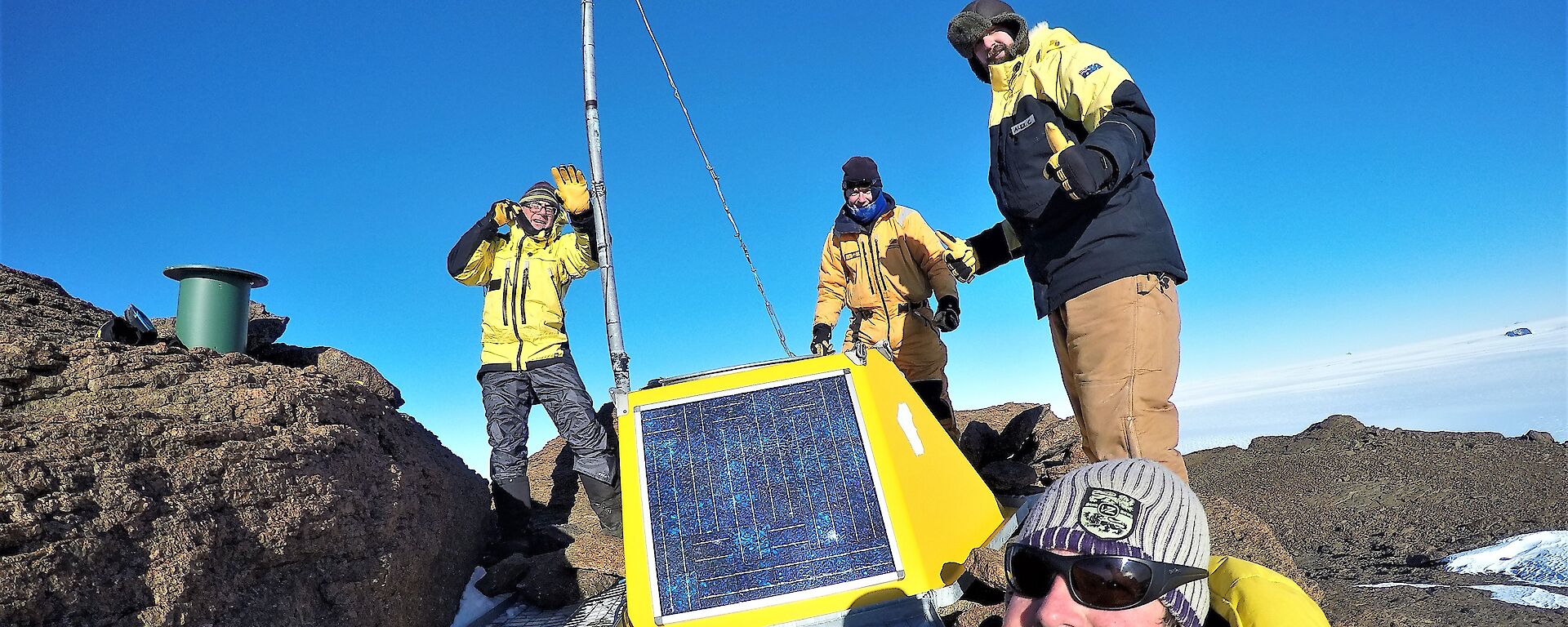 Four men in snow suits stand next to a yellow radio transmitter