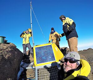 Four men in snow suits stand next to a yellow radio transmitter