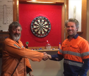 two men stand in front of a dart board shaking hands