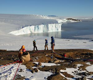 A team of five people stand on a rock edge in harnesses and helmets