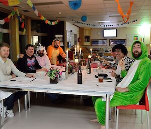 7 men sit around a dinner table, some are dressed in all-in-one animal costumes