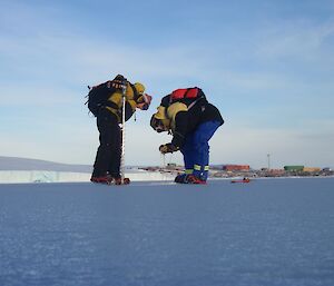 two people crouch on the ice measuring the sea ice with Mawson station in the background