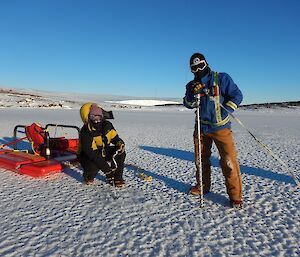 a mns stands holding a sea drill on sea ice and a woman crouches next to him