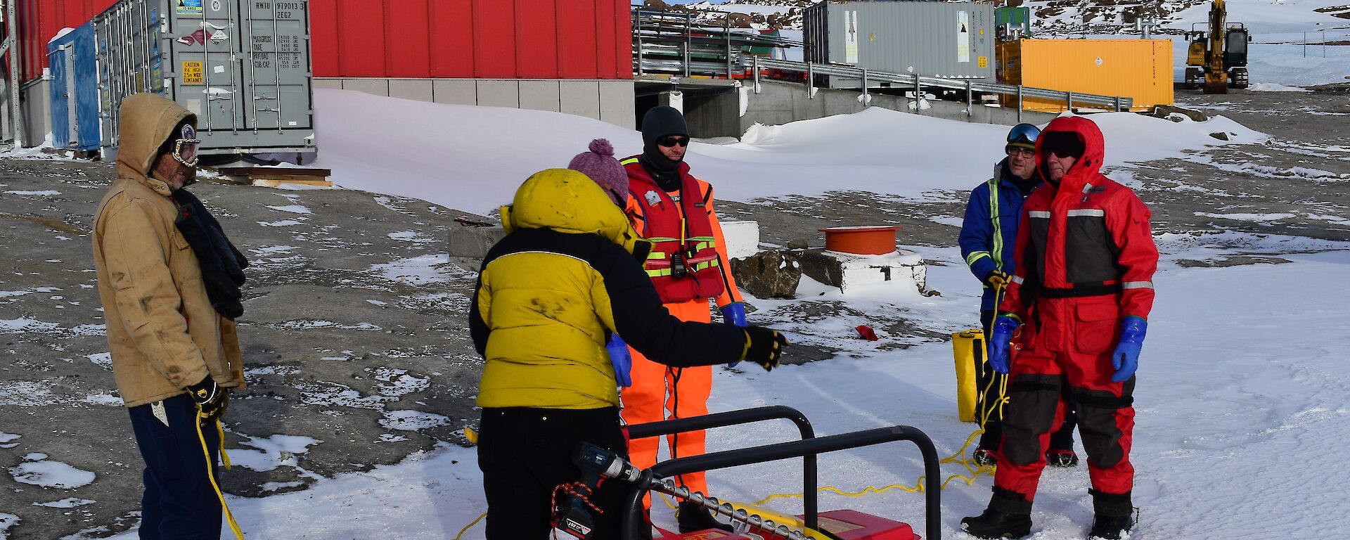 a man in an orange immersion suit stands in front of a rescue platform, while expeditioners prepare the ropes