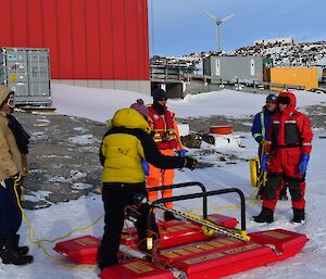 a man in an orange immersion suit stands in front of a rescue platform, while expeditioners prepare the ropes