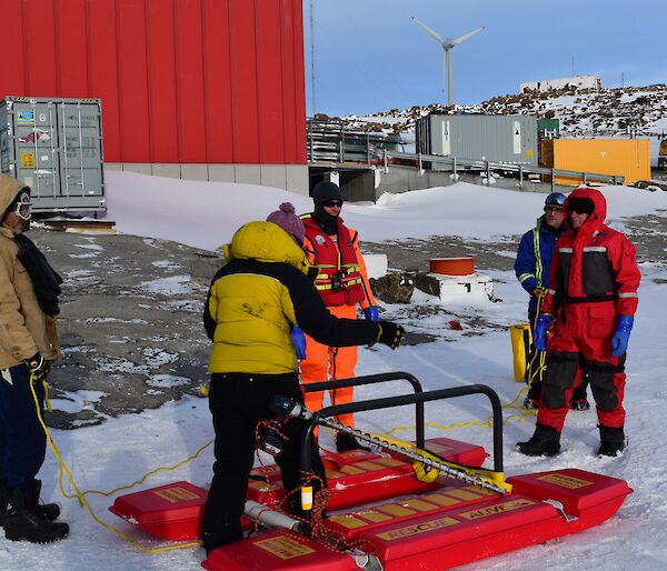 a man in an orange immersion suit stands in front of a rescue platform, while expeditioners prepare the ropes