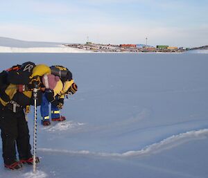two people stand side by side drilling into sea ice with Mawson station in the background