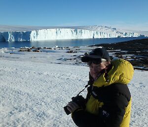 A man in a yellow coat stands in front of a glacier holding a camera