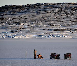 One man crouches the other man stands next to two quad bikes