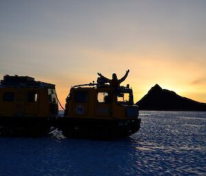 A man stands on a Hägglunds with arms outstretched and the sunrise behind him