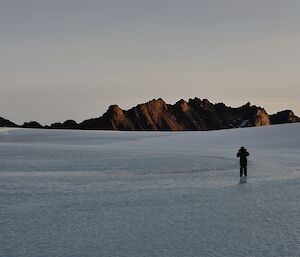A man walks on frozen ice with a mountain range in the background
