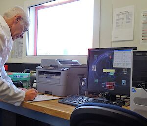 Aa man in a white coat stands in front of a computer writing notes