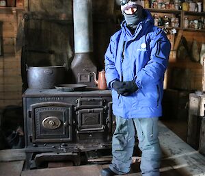 A man in a blue coat and balaclava stands in front of a historic wood fired stove