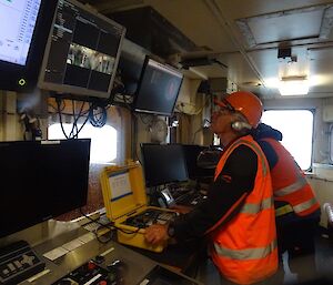 A man in a high visibility jacket and helmet stands in front of a computer screen on a ship