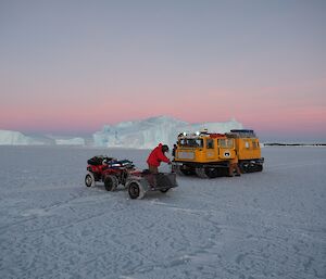 A yellow Hägglunds and red quad bike in front of a pink dawn and icebergs. A man in a red coat is drilling.