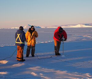 Three men stand on ice with an ice shelf behind them. One is drilling