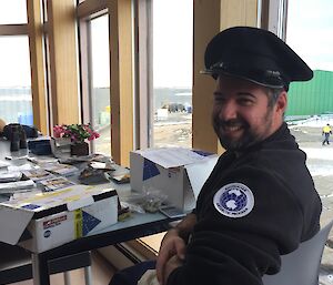 A man sits in front of boxes with a postman’s hat on