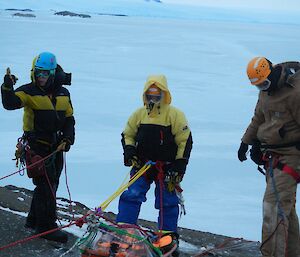 Three people stand on a cliff edge wearing ropes and harnesses