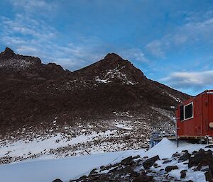 A red hut sits in front of a mountain range and snow covered valley
