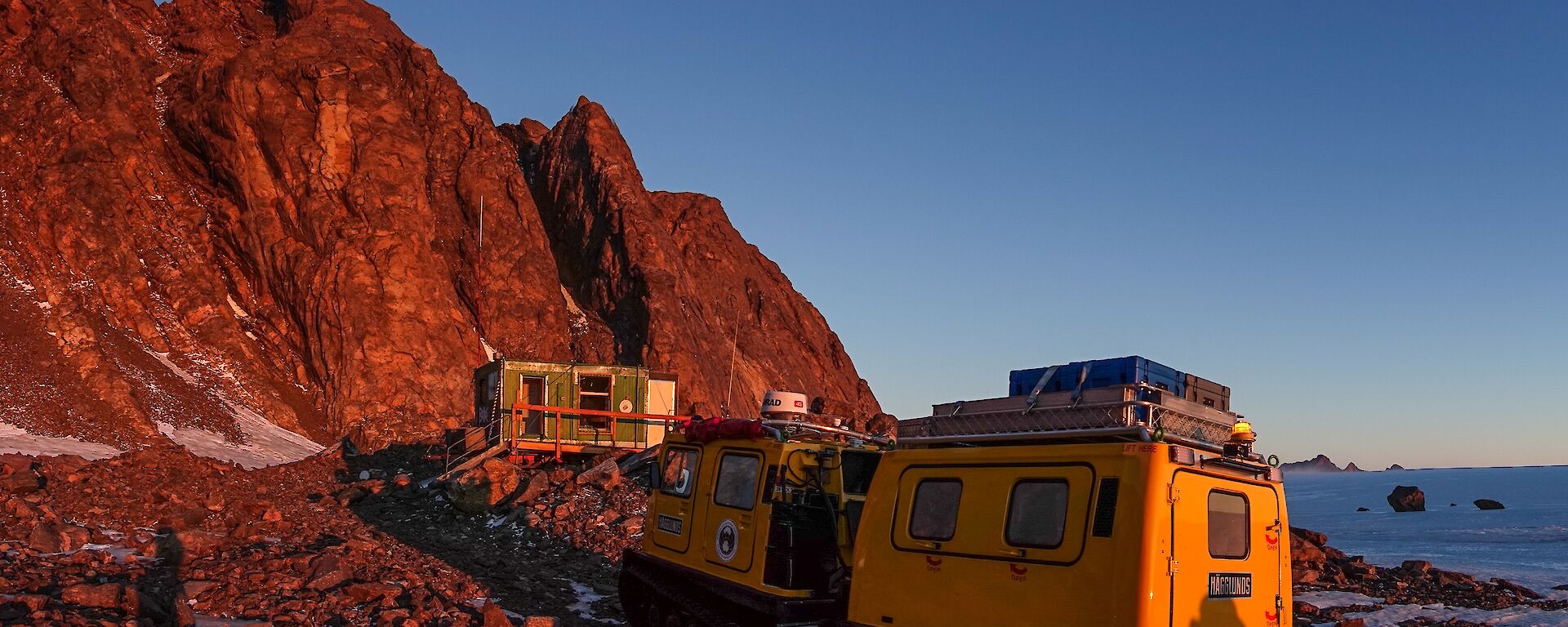 A yellow Hägglunds is parked in front of a green hut positioned in front of mountains