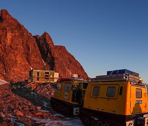 A yellow Hägglunds is parked in front of a green hut positioned in front of mountains