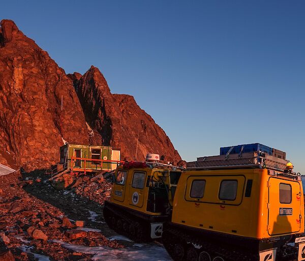 A yellow Hägglunds is parked in front of a green hut positioned in front of mountains