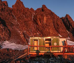 A green field hut with no windows or doors sits in front of a mountain peak