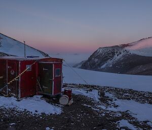 A little red hut in front of a mountain plateau and pink sky