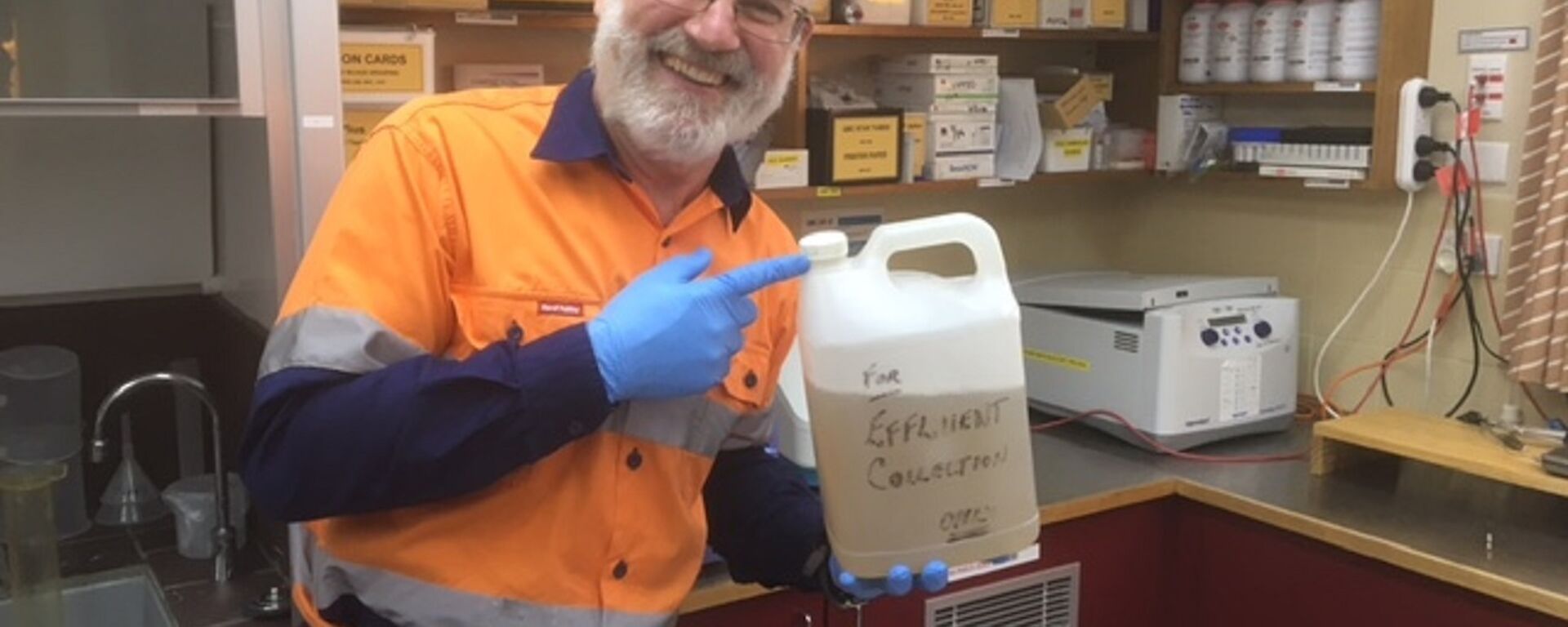 A man in high visibility clothing stands in a laboratory holding a container of liquid