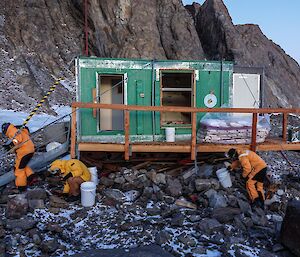 Three people in yellow freezer suits are collecting in front of a hut