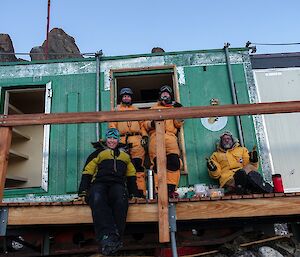 Four people in yellow freezer suits sit on the deck of a hut