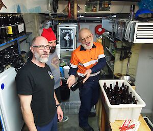 Three men stand in a brewery in front of beer bottles