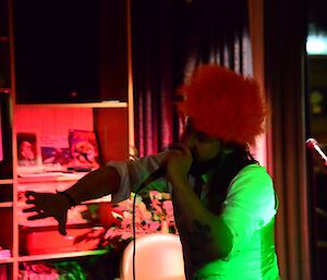 A man stands in an orange wig with a microphone in his hand