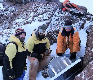 Three men stand next to a radio repeater on a mountain.