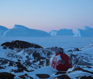 A red dome shaped hut sits amongst ice covered rocks and icebergs