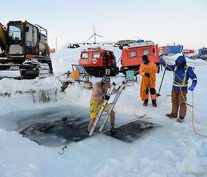 A man in yellow shorts descends a ladder into a frozen pool Surrounded by machinery and men in warm clothing