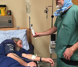 A man sleeps in a hospital bed with a man in theatre scrubs attending to him.