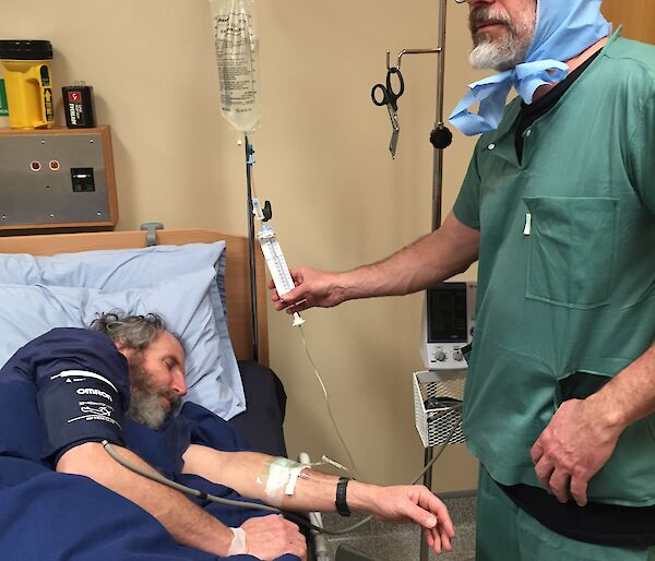 A man sleeps in a hospital bed with a man in theatre scrubs attending to him.