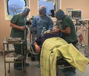 Three men in theatre scrubs attend to a patient on a hospital bed.