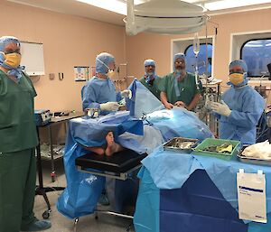 Five men in theatre scrubs and masks stand around a patient in a theatre