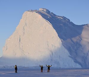 Three expeditioners stand in front of a pyramid shaped ice berg