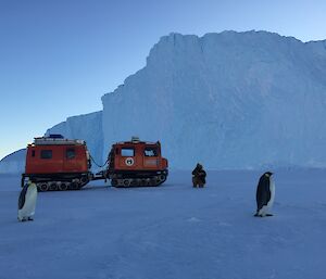 An orange Hägglunds in front of an iceberg with emperor penguin