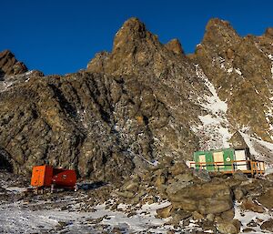 An orange caravan next to a green field hut at the base of a mountain range