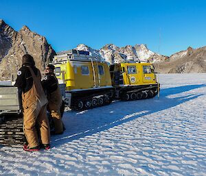 Two men stand next to a trailer on the sea ice in front of a mountain range